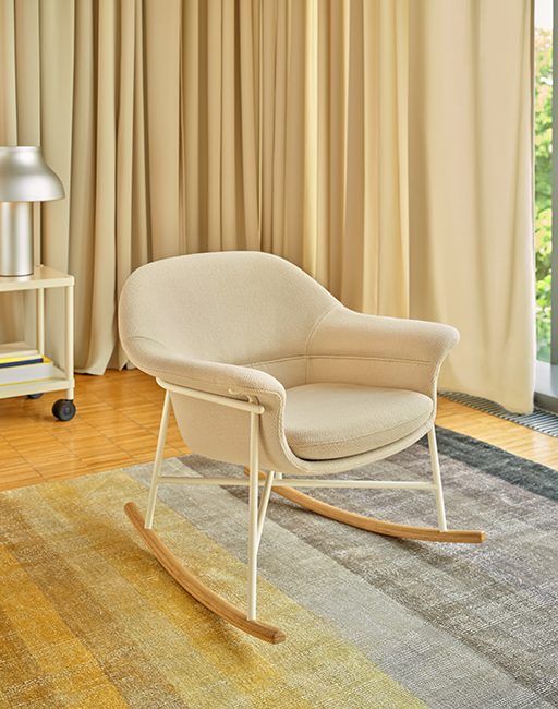 A light-coloured rocking chair with wooden runners placed on a carpet in a spacious interior with a large window