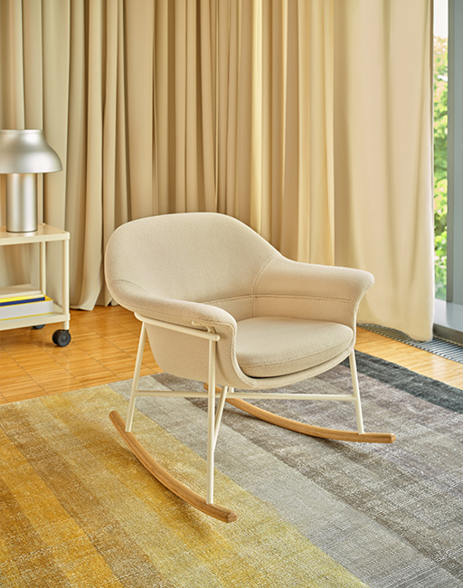  A light-coloured rocking chair with wooden runners placed on a carpet in a spacious interior with a large window